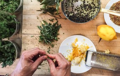 hands pinching at herbs next to a plate with lemon zest and ingredients in a pot