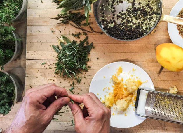 hands pinching at herbs next to a plate with lemon zest and ingredients in a pot