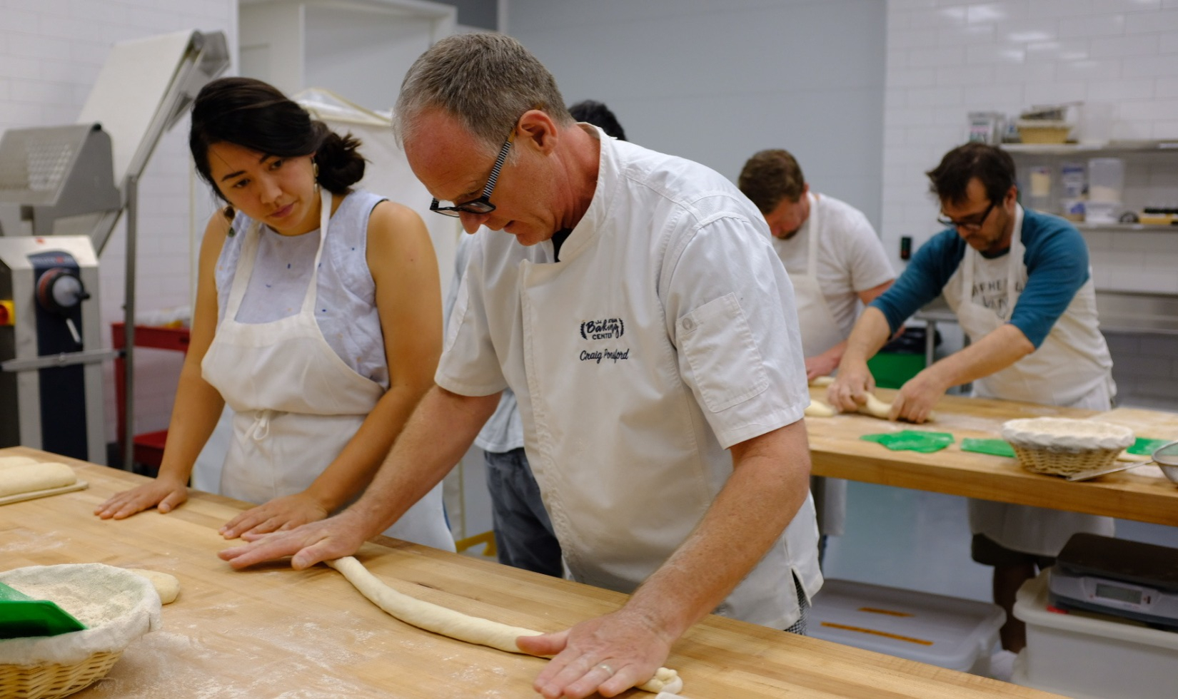 Woman in cooking classes watches instructor roll pastry on butcher block table.