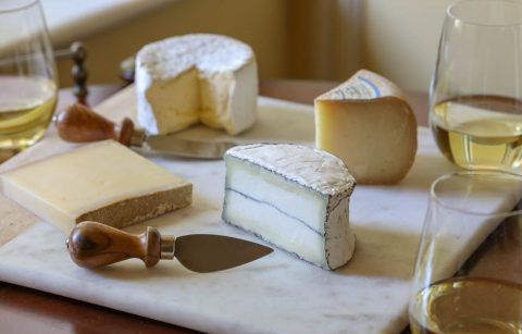 four different types of cheese on a cheese board next to poured glasses of Chardonnay