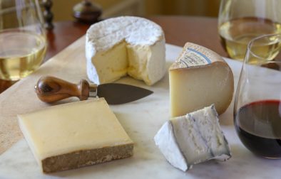 four different types of cheese on a cheese board next to poured glasses of wine