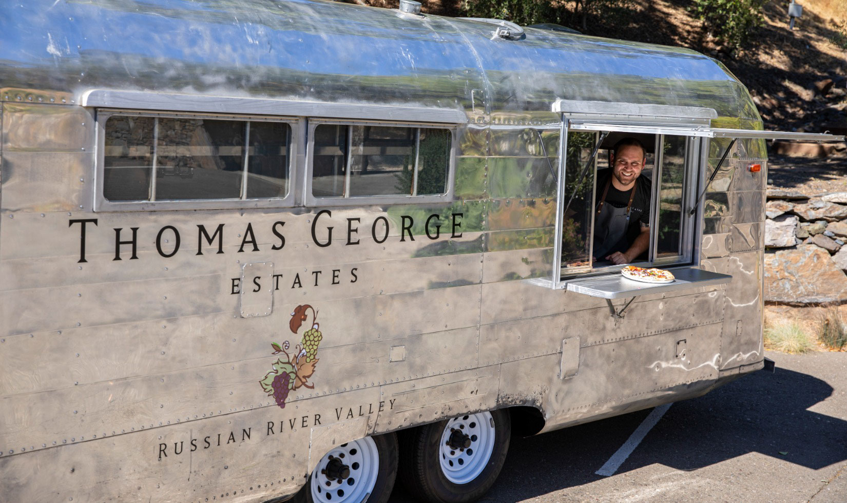 Outdoor Wine Tastings A silver airstream with the thomas george logo with a window open serving pizza