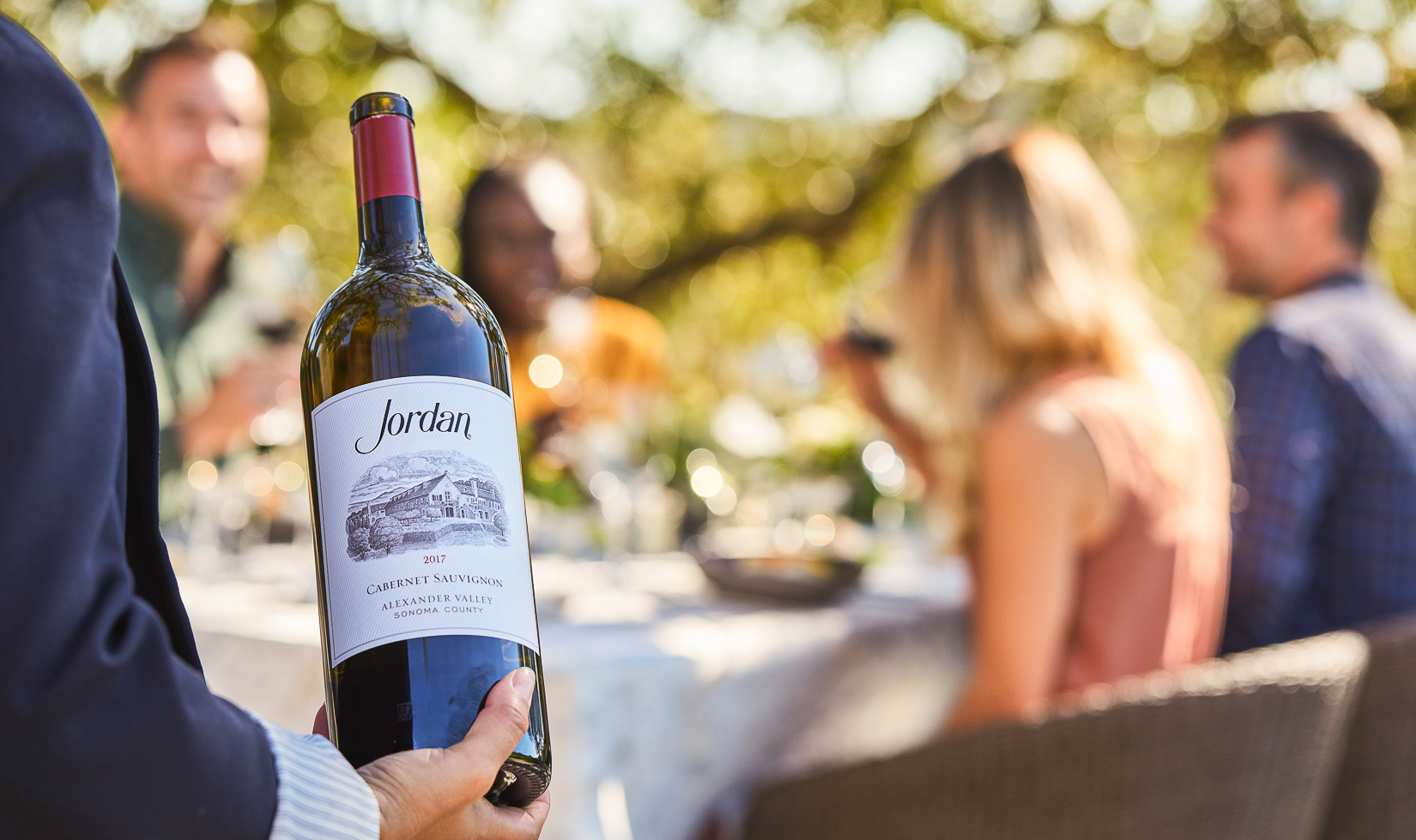 woman holding a large bottle of cabernet sauvignon in focus with a table of four guests in the background