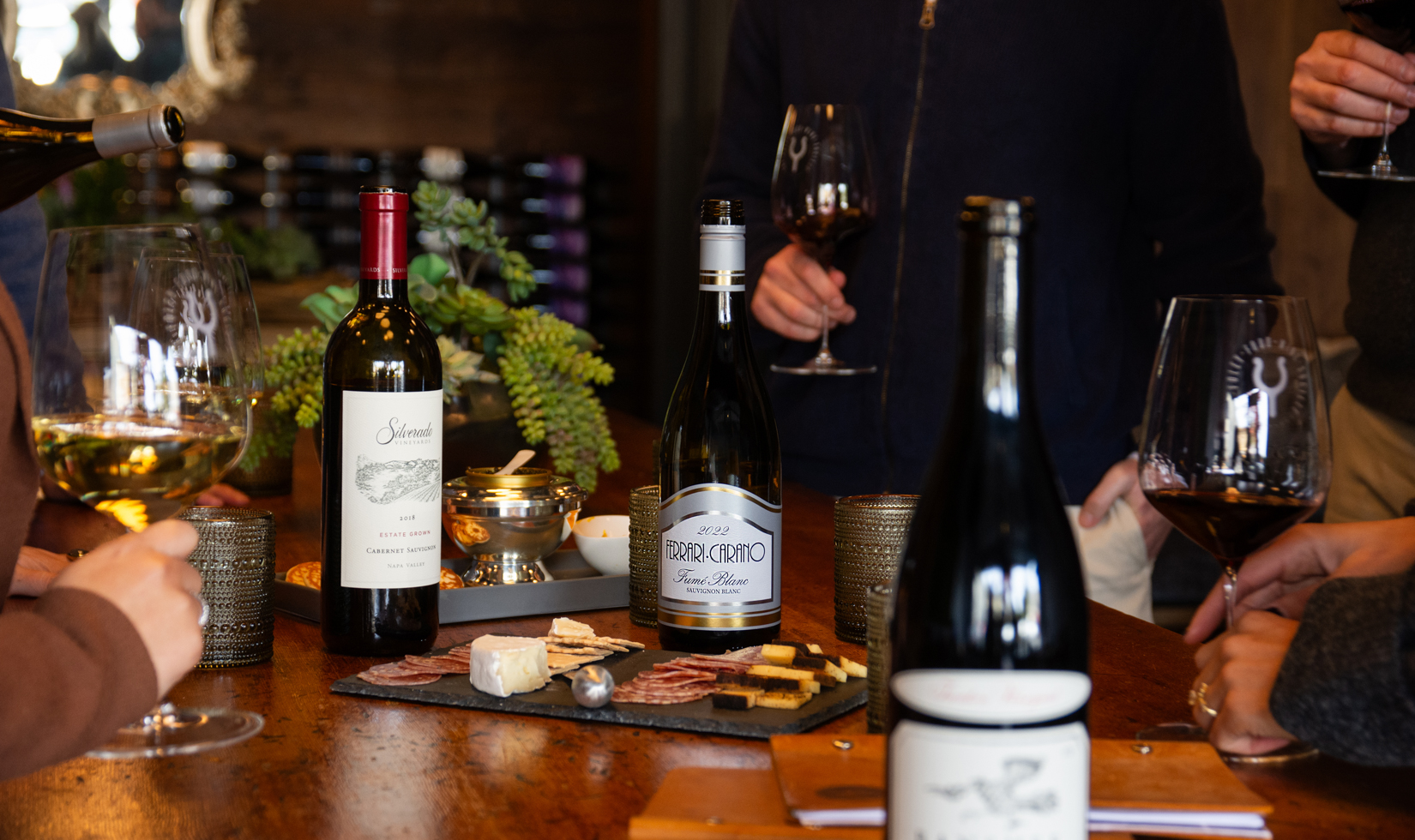 people gathered around table with bottles of wine and a cheese plate