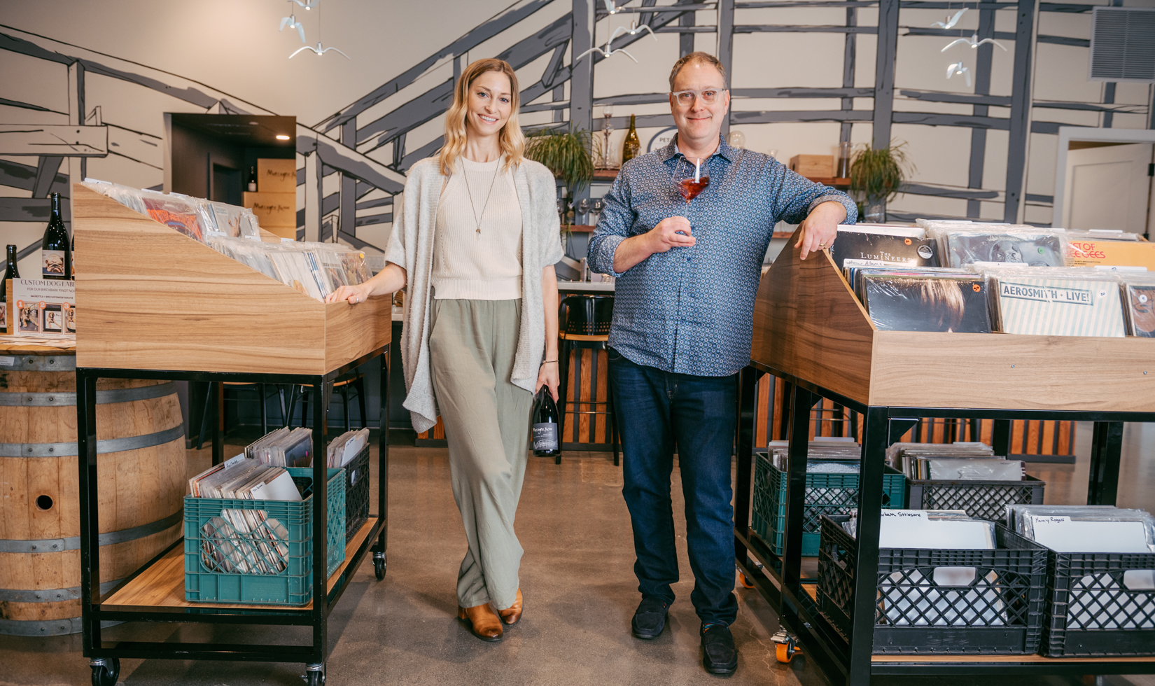 a man and a woman standing in a vinyl music shop holding a glass of wine and bottle of wine