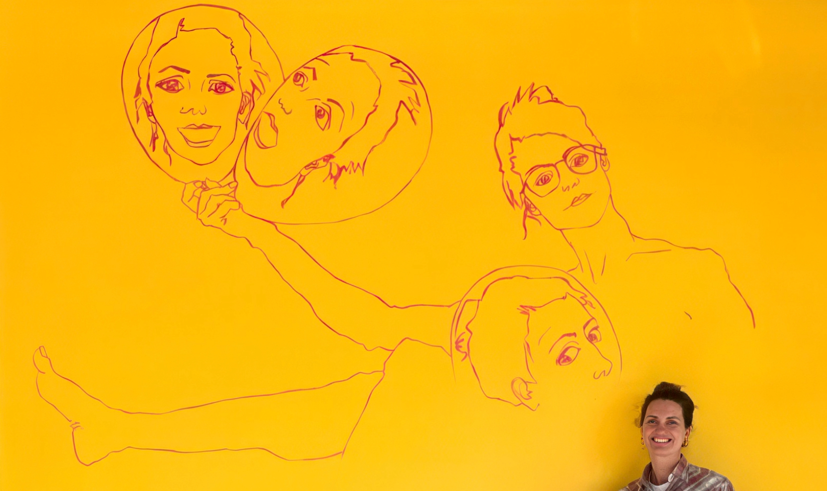 Artwork featuring figurative drawings on a bright yellow wall with the artist sitting beneath the drawing. 