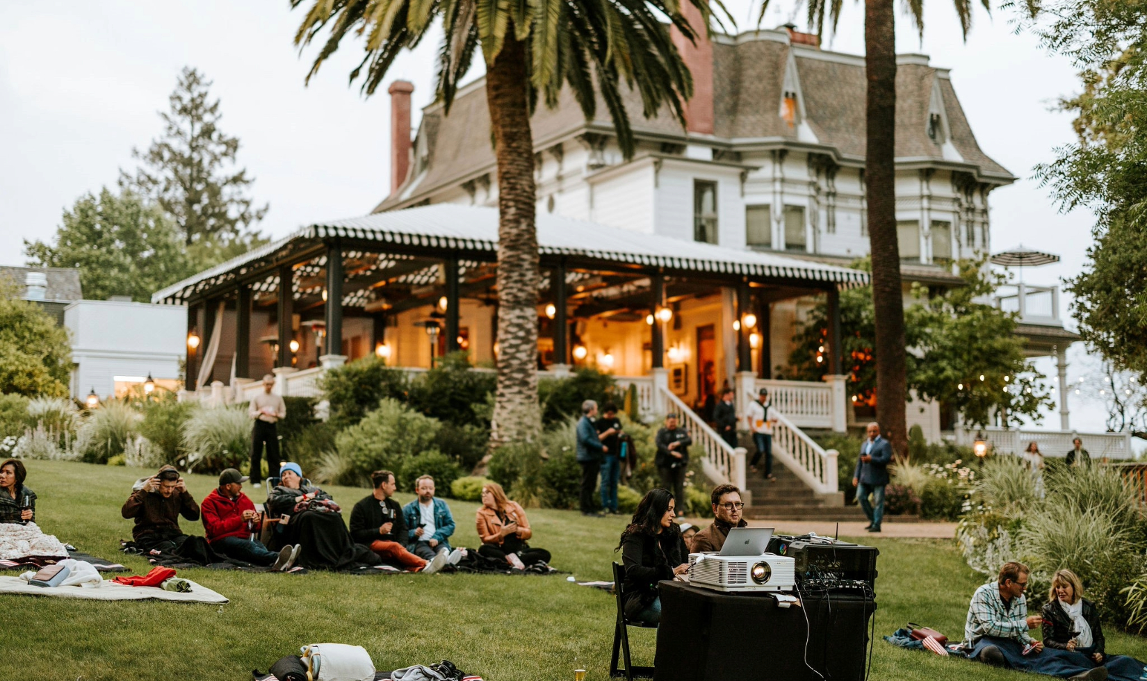 groups sitting in blankets on grass lawn in front of victorian inn watching movies projected onto an outdoor screen