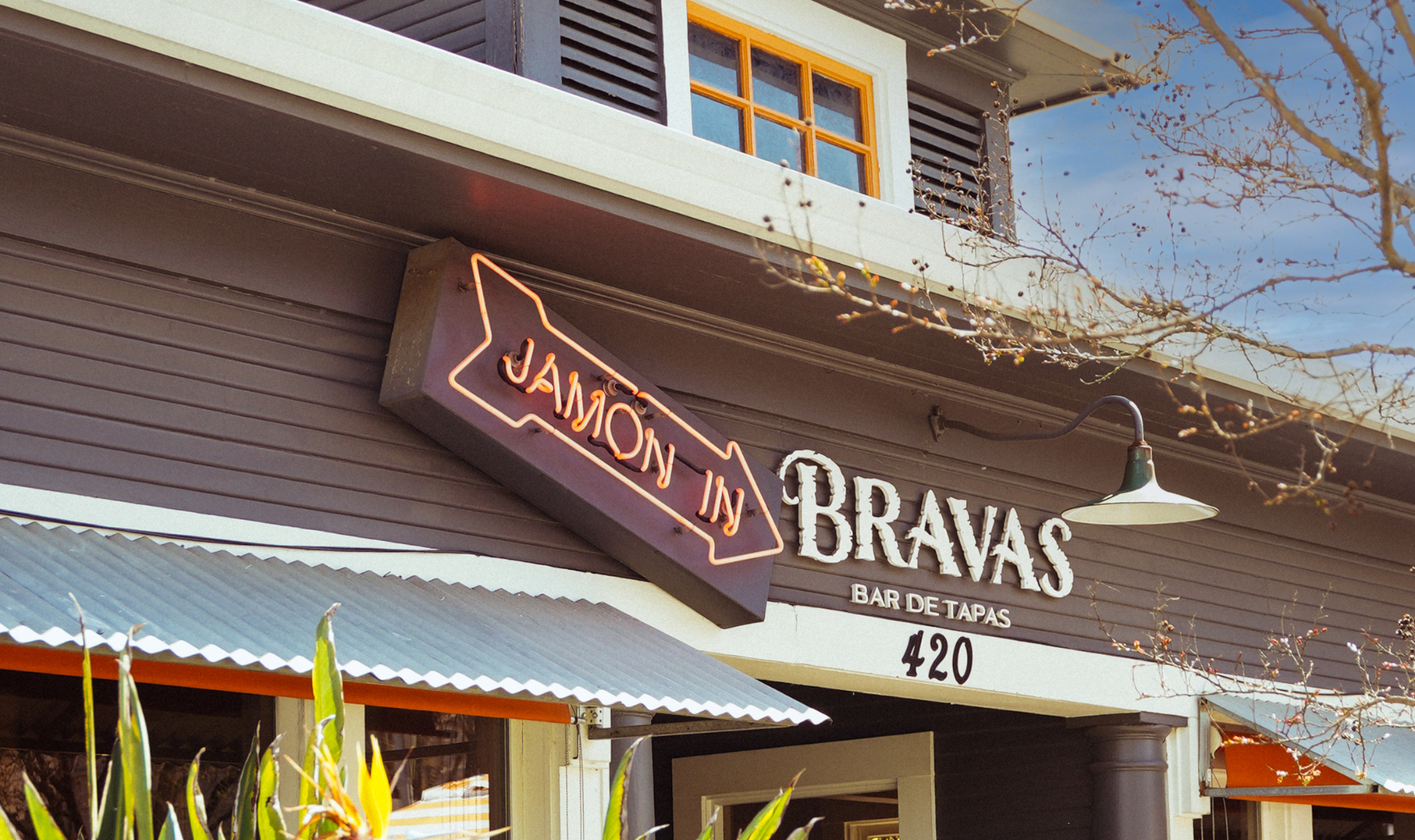 restaurant exterior with neon sign over entrance door that read "jamon in" next to a wood sign that reads "bravas bar de tapas"
