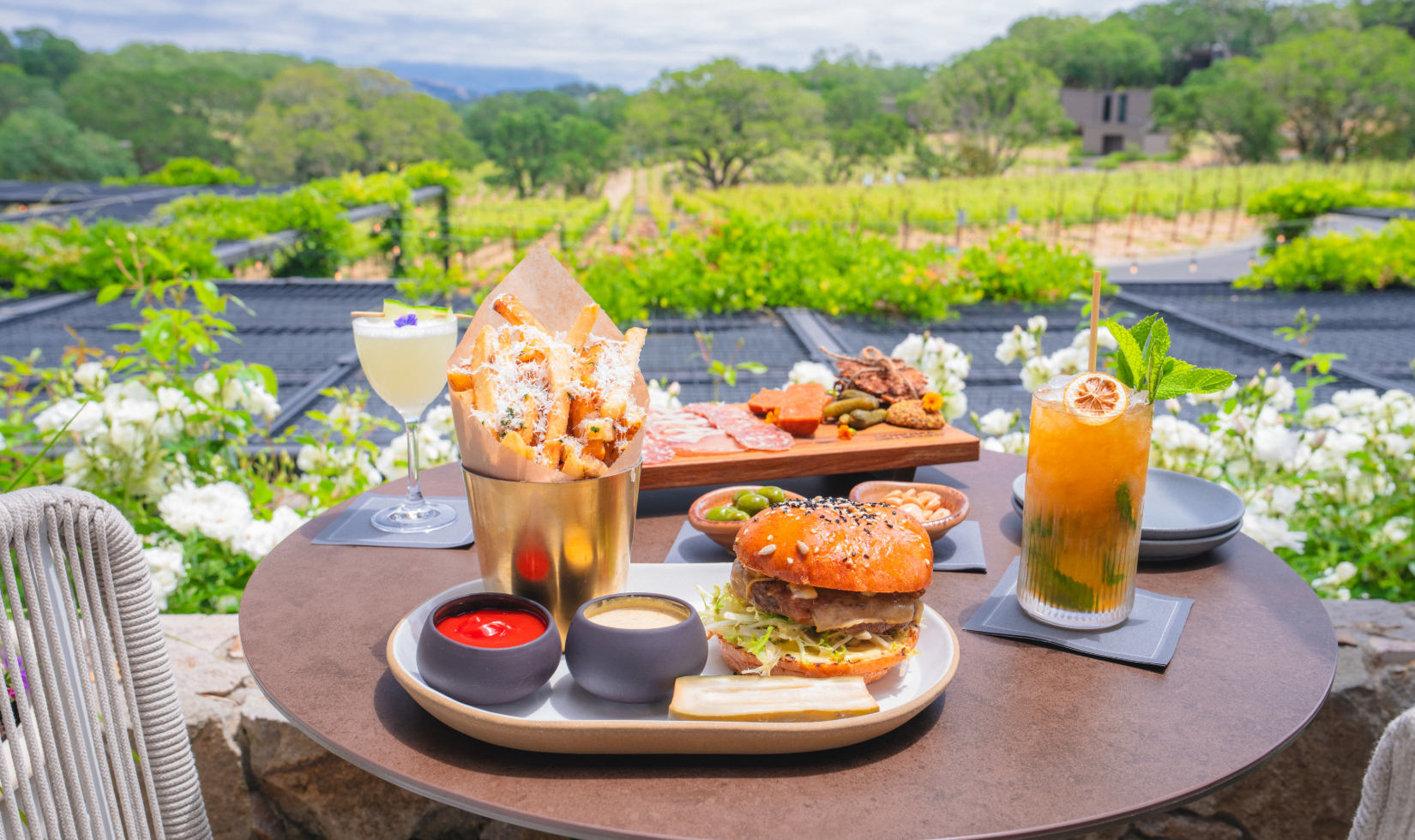 outdoor restaurant table with cheeseburger, fries, cocktails and charcuterie board overlooking view of vineyards and resort grounds