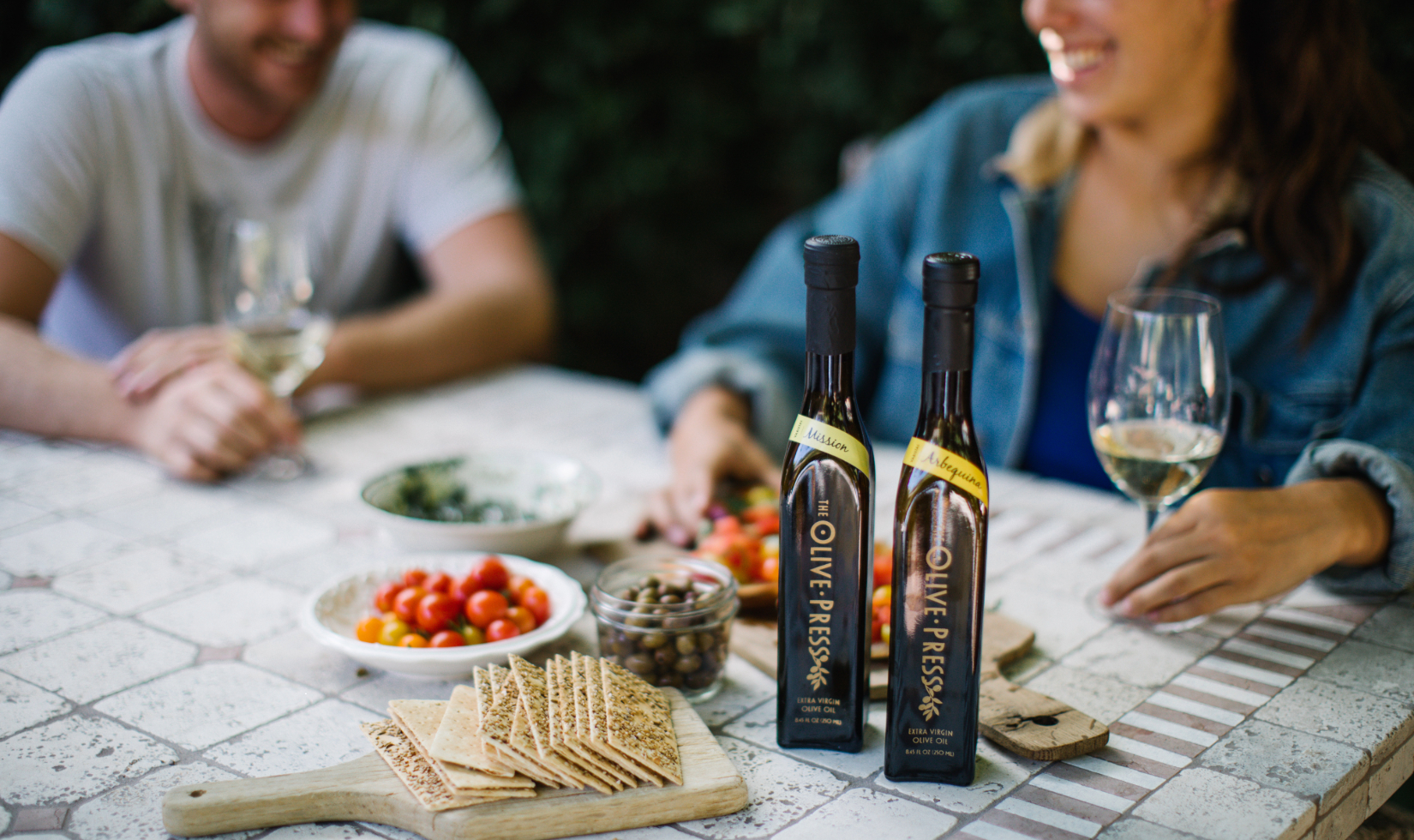 Two people seated at table set up for an olive oil tasting with crudité and crackers.