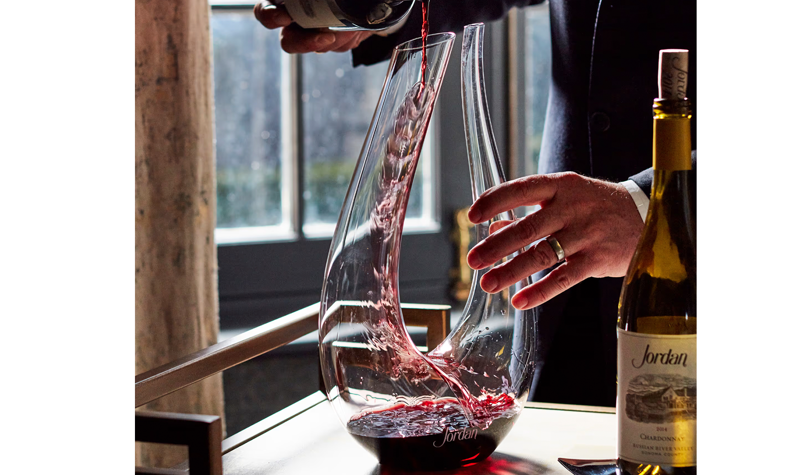 Red wine being poured into a Jordan-branded decanter with a bottle of Jordan Chardonnay nearby.